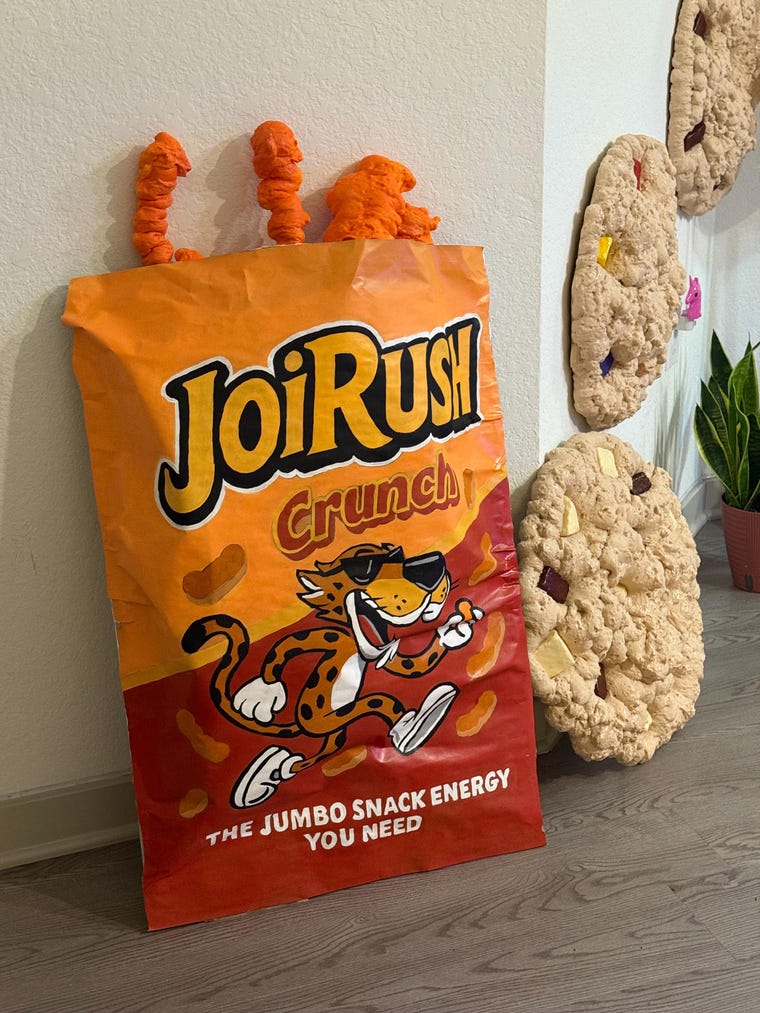 Oversized JOIRUSH Crunch snack bag sculpture leaning beside jumbo cookie wall art in the studio